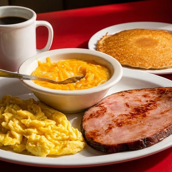 A breakfast plate with scrambled eggs, ham, cheesy grits, pancakes, and a cup of coffee.