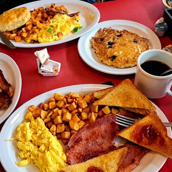 A red table laden with multiple plates of breakfast food, including eggs, bacon, toast, and pancakes.