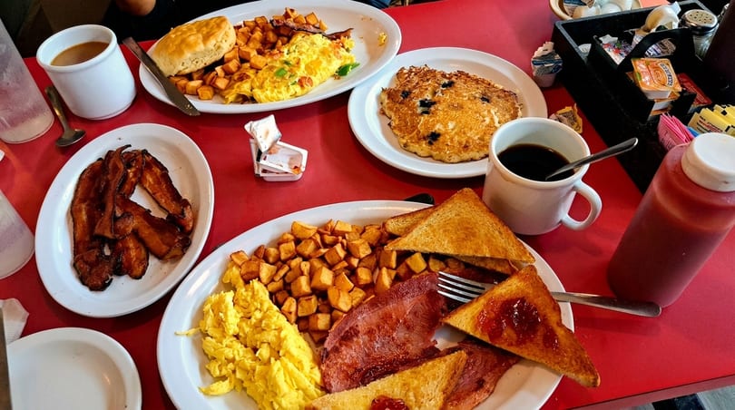 A table laden with multiple plates of breakfast food, including eggs, bacon, toast, and pancakes.